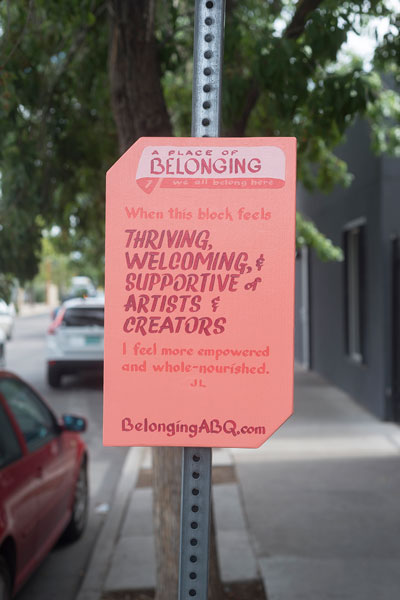 Sign reads, "A place of belonging #1, We all belong here. When this block feels thriving, welcoming, and supportive of artists and creators, I feel more empowered and whole-nourished. JL BelongingABQ.com." Salmon colored sign on a post on a sidewalk with trees and parked cars in the background.