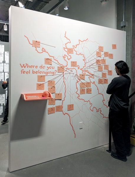 Wall painted with map of bay area in salmon colored paint, with the text "Where do you feel belonging?" There's cards on the map with yarn and pins to different locations. There's a pink shelf with yard, scissors and pins. A visitor is close, reading the cards.