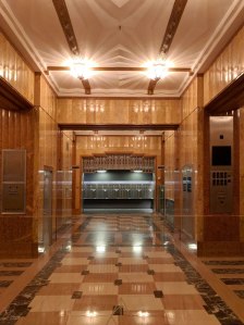 An ornate, gold, orange and red interior. Lots of embellishment on every surface. The ceiling has built-in light figures that make geometric rays of light due to the bas relief in the ceiling. The walls have a matching louvered paneling in gold. There are multiple spaces defined by transoms with ornate floral grillwork. The floors have a checkerboard of yellow and cream marble with black and white marble interspersed and used as a border. There are touches of gleaming metal.