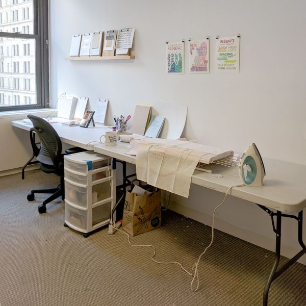 a office with carpet and two folding tables side by side, with an office chair. there's a small ironing board with a bandana on top, and an iron. There are drawings on the wall, and various clipboards, pencils, colored pencils, etc on the table.