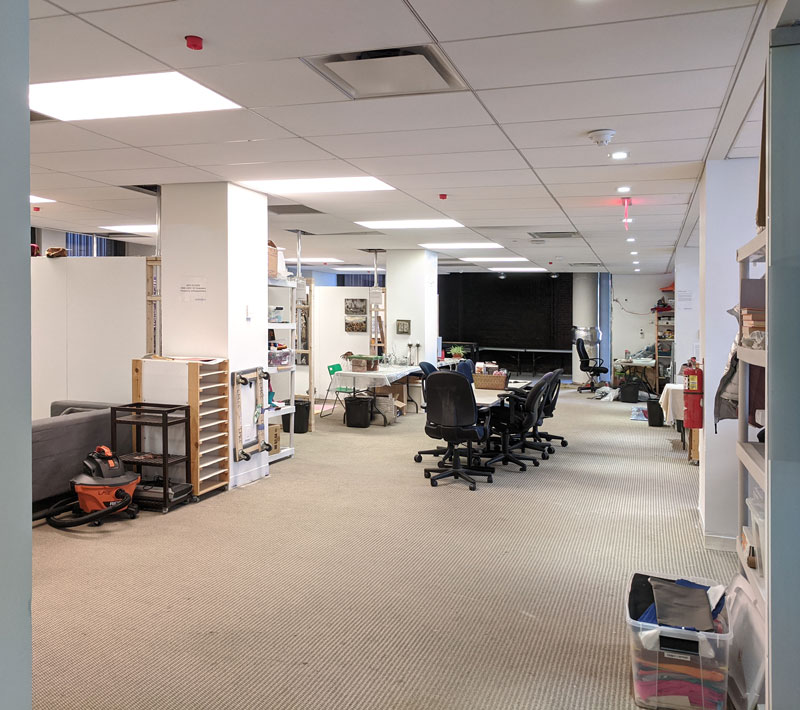 Carpeted office space divided into artist studios with office chairs around a table in the middle. Various art studio supplies: shop vacuum, bin of fabrics, dolly, etc around.