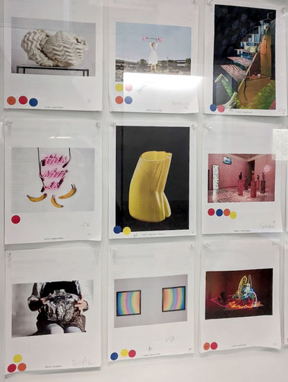 A window case behind glass of nine printouts, each bearing a photo of glass art, with colored dot stickers below.