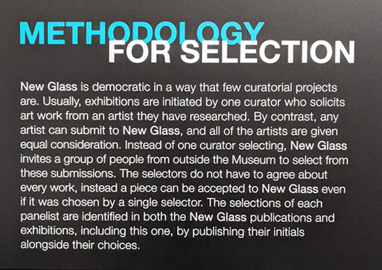 Signage that states, "Methodology for Selection. New Glass is democratic in a way that few curatorial projects are. Usually, exhibitions are initiated by one curator who solicits art work from an artist they have researched. By contrast, any artist can submit to New Glass, and all of the artists are given equal consideration. Instead of one curator selecting, New Glass invites a group of people from outside the Museum to select from these submissions. The selectors do not have to agree about every work, instead a piece can be accepted to New Glass even if it was chosen by a single selector. The selections of each panelist are identified in both the New Glass publications and exhibitions, including this one, by publishing their initials alongside their choices."