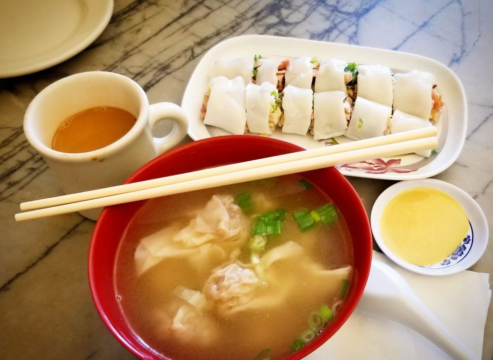 Photo of a red bowl with broth and wontons. A mug with possibly HK style milk tea, and a rice roll with a side of mustard.