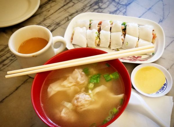 Photo of a red bowl with broth and wontons. A mug with possibly HK style milk tea, and a rice roll with a side of mustard.