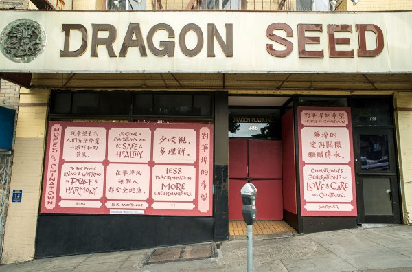 Photo of Dragon Seed Bridal and Photography storefront. A big sign above reads, "Dragon Seed" in brown text on white background. Below, the window is boarded up and covered in a wheatpasted poster. The text on the poster is in English and Chinese. It reads: Hopes for Chinatown. To see people living and working in peace and harmony, by Alina. Everyone in Chinatown will be safe and healthy. Anonymous. Less discrimination. More Understanding. YY. Chinatown's Generations of love and care will continue. Sunflower. The text is in red in light pink boxes on a background of red with a scale-like pattern of overlapping concentric circles.