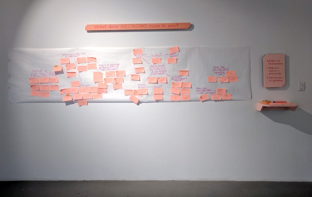 A photo of a gallery wall with a piece of paper with questions about belonging in it. There are also salmon pink signs that say "What does belonging mean to you?"