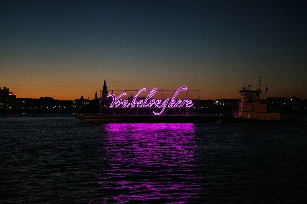 a photo of a neon installation with the text, 'You belong here' in puruple script.' the neon is installed on a waterfront, so it reflects in the water. The photo was taken after sunset.